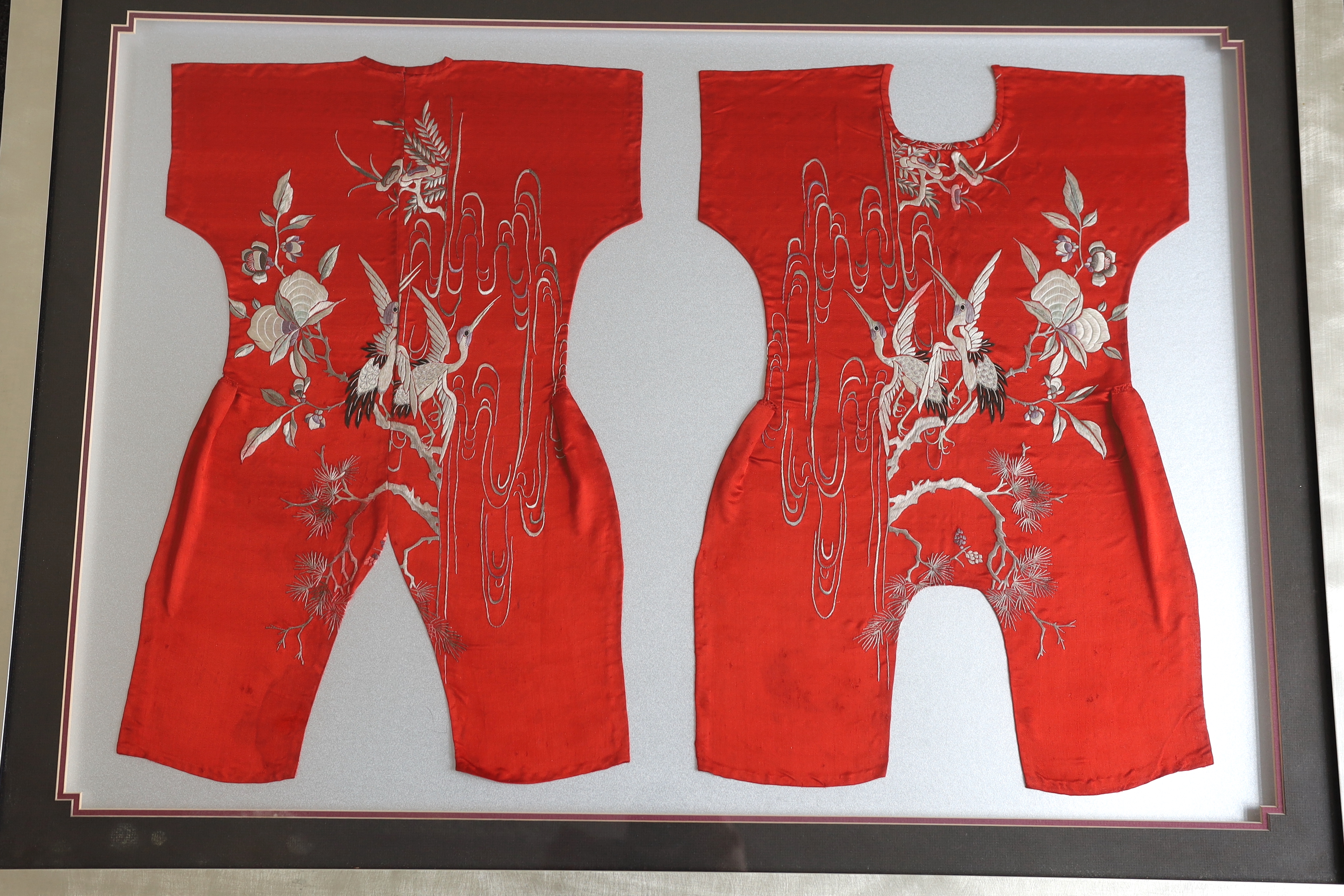 Three early 20th century Chinese framed embroideries: two purple baby’s bibs embroidered with multi-coloured silk threads, and another red silk child’s “romper suit”, embroidered with white and pastel embroidery, mounted